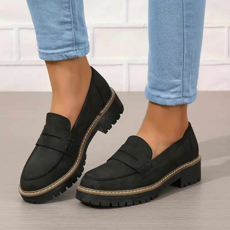 Sorte loafers med tyk tandet sål og kontrastsyning, båret med oprullede lyseblå jeans foran hvid murstensvæg. 6