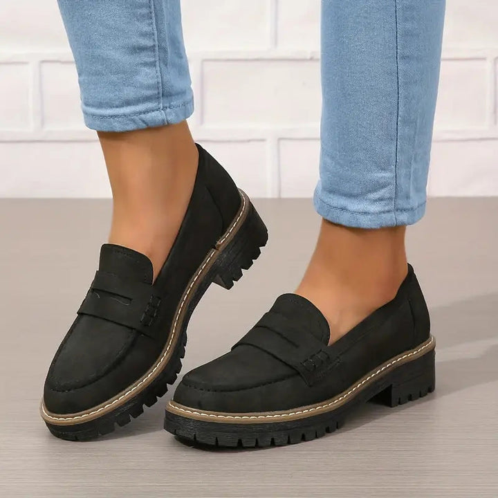 Sorte loafers med tyk tandet sål og kontrastsyning, båret med oprullede lyseblå jeans foran hvid murstensvæg. 6