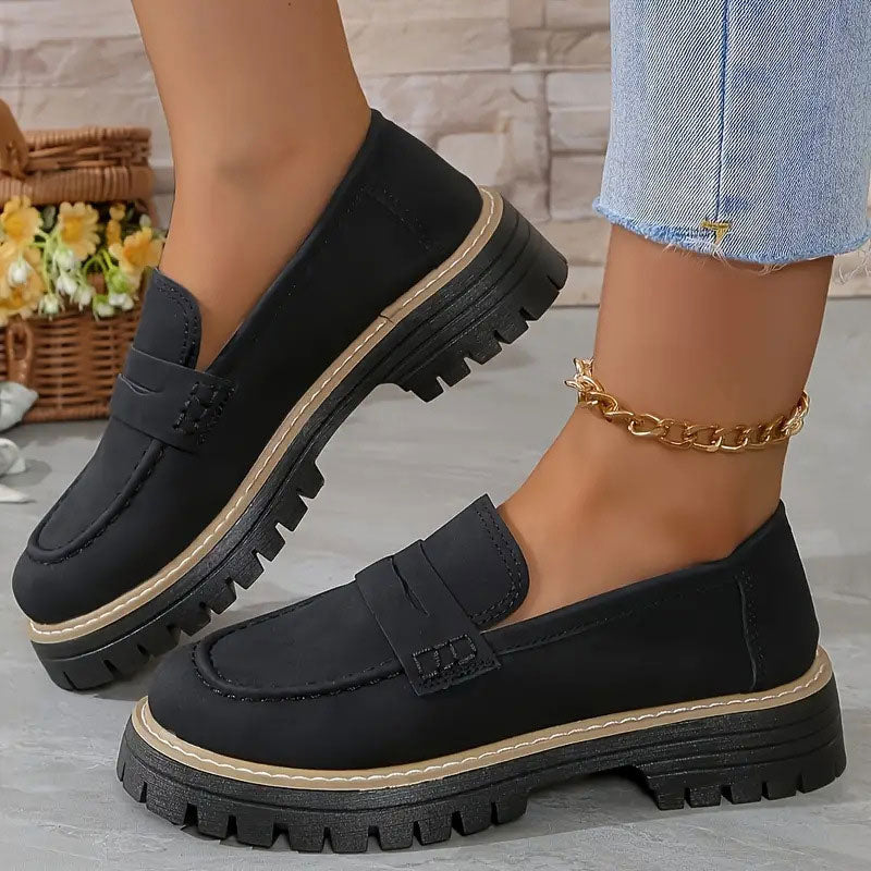 Sorte loafers med kraftig platformsål og beige kant, båret med guldfarvet ankelkæde ved siden af kurv med blomster. 12