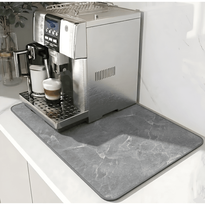 Grå måtte med marmormønster og mørkere kant under en kaffemaskine på en hvid bordplade. 12