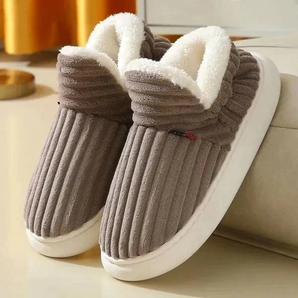 Par riflede slippers i gråbrun med hvid plysforing og tyk hvid sål, stillet på et lyst gulv ved en sofa. 3